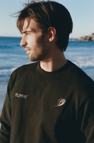 A man wearing a black organic cotton sweater with surf logo with sea in the background